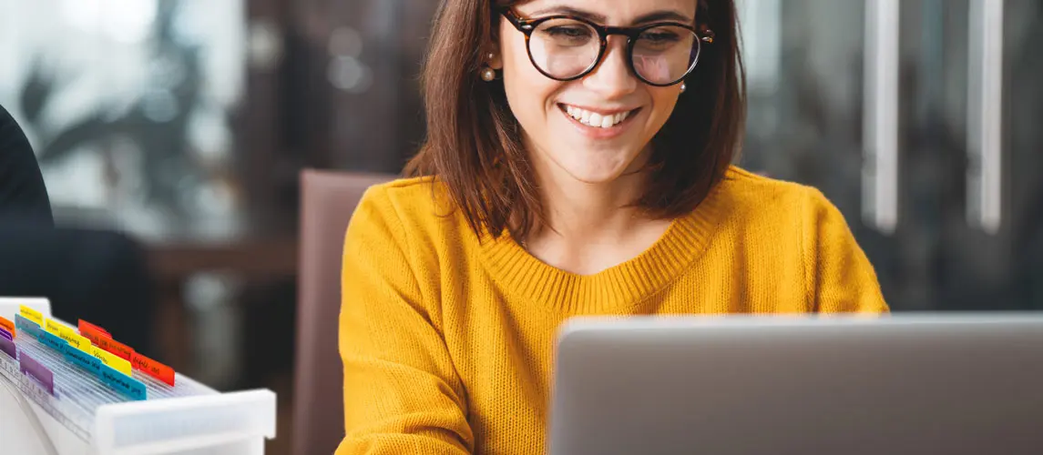 Strukturiertes Arbeiten Eine lächelnde Frau mit Brille und gelbem Pullover arbeitet konzentriert an einem Laptop im Büro, neben ihr ein transparentes Ablagesystem mit farbigen Registern von MAPPEI, für eine organisierte Arbeitsweise.