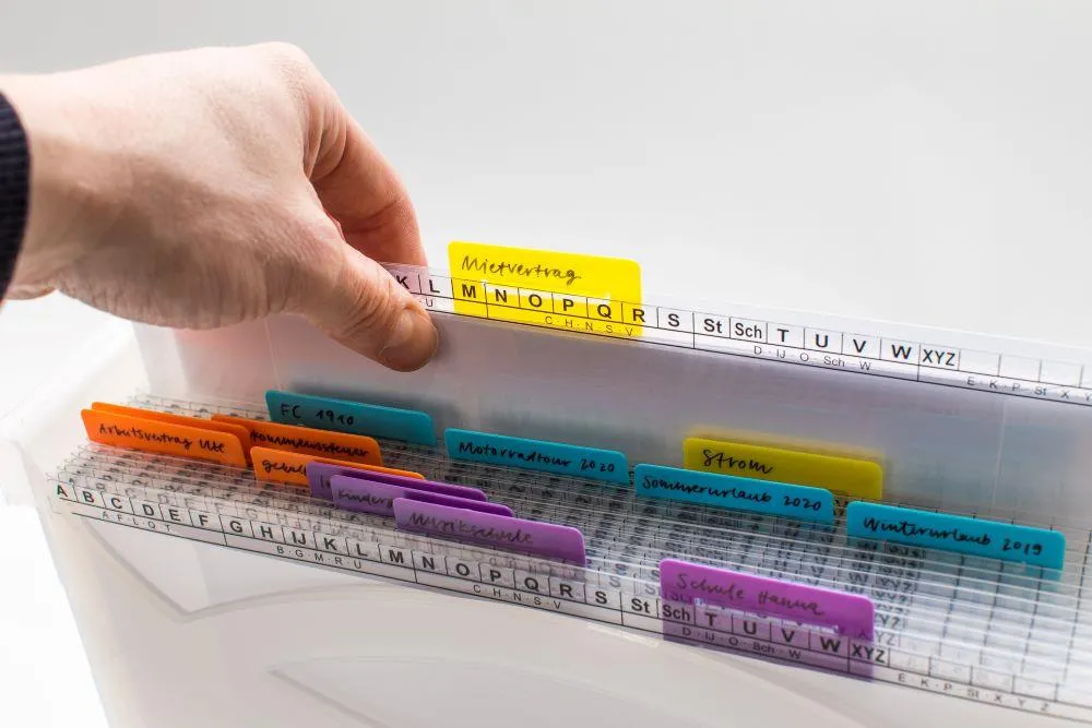 Nahaufnahme einer Ordnungsbox gefüllt mit durchsichtigen Aktionsmappen, am oberen Rand ist eine Ordnungsleiste aufgedruckt, oben an den Mappen sind verschiedenfarbige Beschriftungsläufer angebracht, eine Hand zieht ein Mappe aus der Box heraus