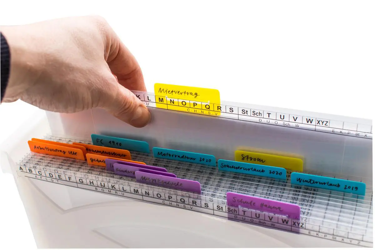 Eine Hand greift in eine transparente MAPPEI Hängebox mit farbigen Registern, die verschiedene beschriftete Reiter für eine übersichtliche Ablage persönlicher Dokumente wie "Mietvertrag", "Arbeitsvertrag" und "Urlaub" enthält.