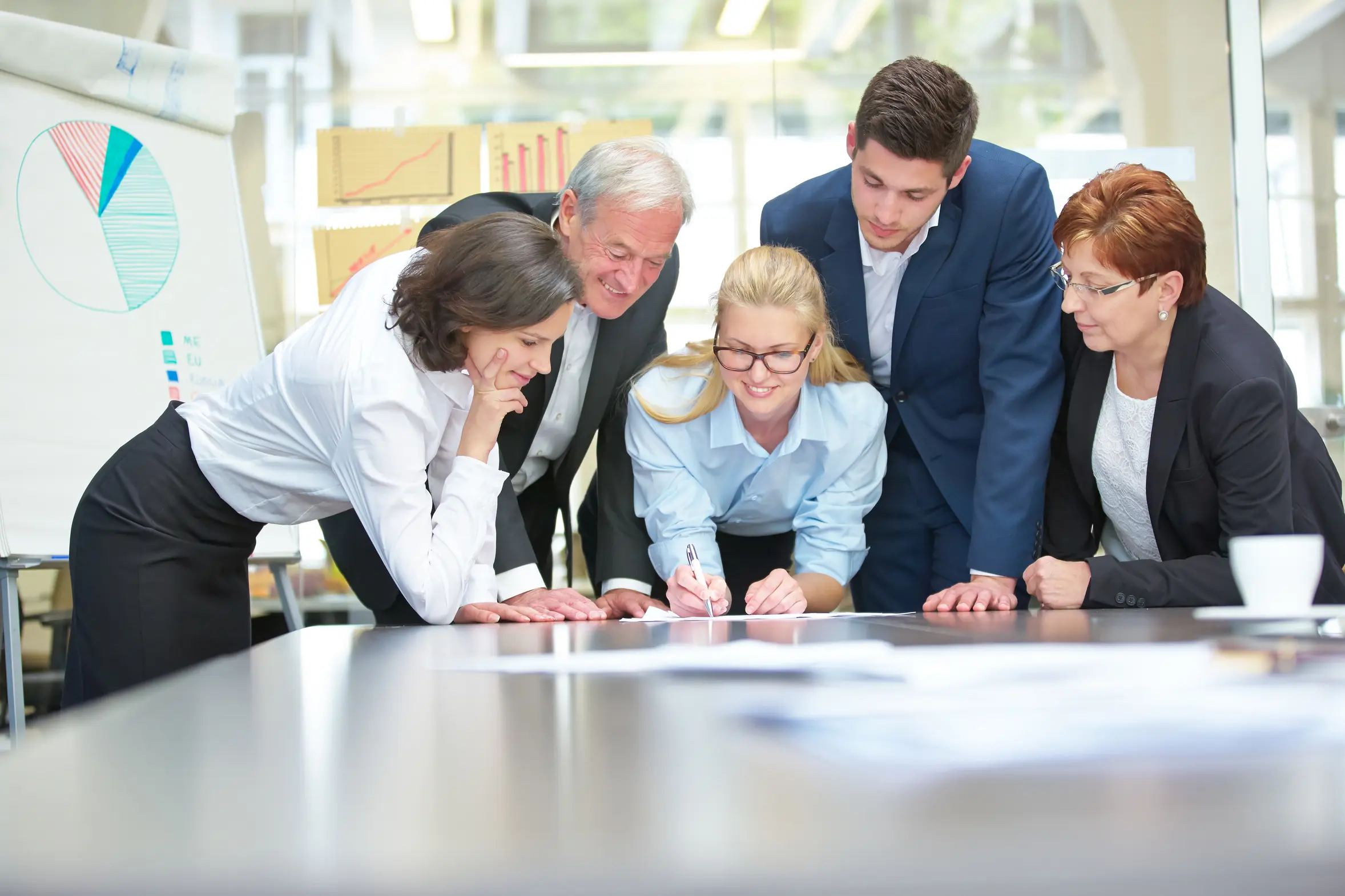 Fünf Personen in Business-Kleidung beugen sich über einen großen Tisch in einem hellen Besprechungsraum und betrachten Dokumente.