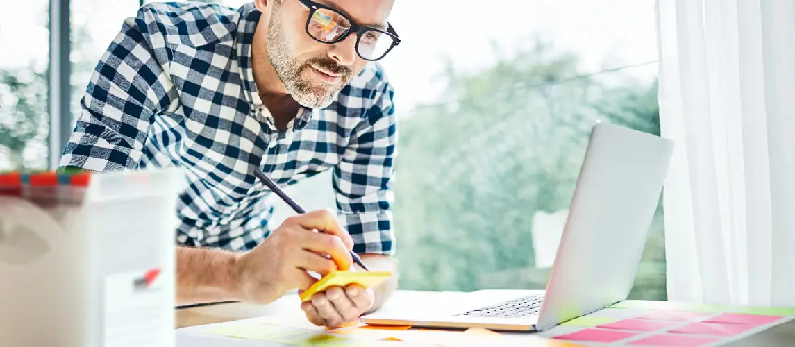 Mann im Homeoffice Mann mit Brille und kariertem Hemd notiert Ideen auf einem gelben Haftnotiz am Schreibtisch mit Laptop und Büromaterialien für effizientes Arbeiten.