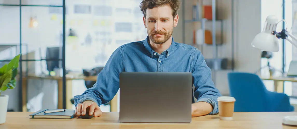 Homeoffice mit Mappei Mann mit Bart und blauem Hemd arbeitet konzentriert an einem Laptop in einem modernen Homeoffice mit Notizbuch, Stift und Kaffeetasse auf einem hellen Holztisch.