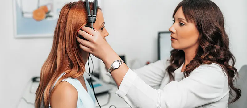 Eine junge Frau mit langen roten Haaren erhält einen Hörtest von einer Hörgeräteakustikerin mit braunen Haaren und weißem Kittel, die ihr Kopfhörer anlegt, in einem Untersuchungsraum.