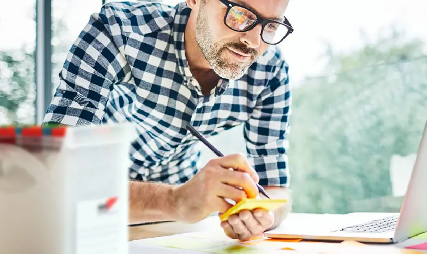 Mann mit Brille und kariertem Hemd notiert Ideen auf einem gelben Haftnotiz am Schreibtisch mit Laptop und Büromaterialien für effizientes Arbeiten.