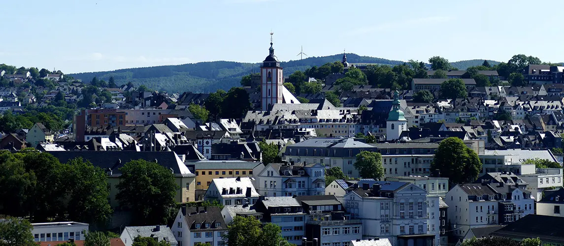 Panoramablick auf Siegen, Deutschland, mit der markanten Martinikirche im Zentrum, umgeben von dicht bebauten Wohngebieten und grünen Hügeln unter einem hellen Himmel.