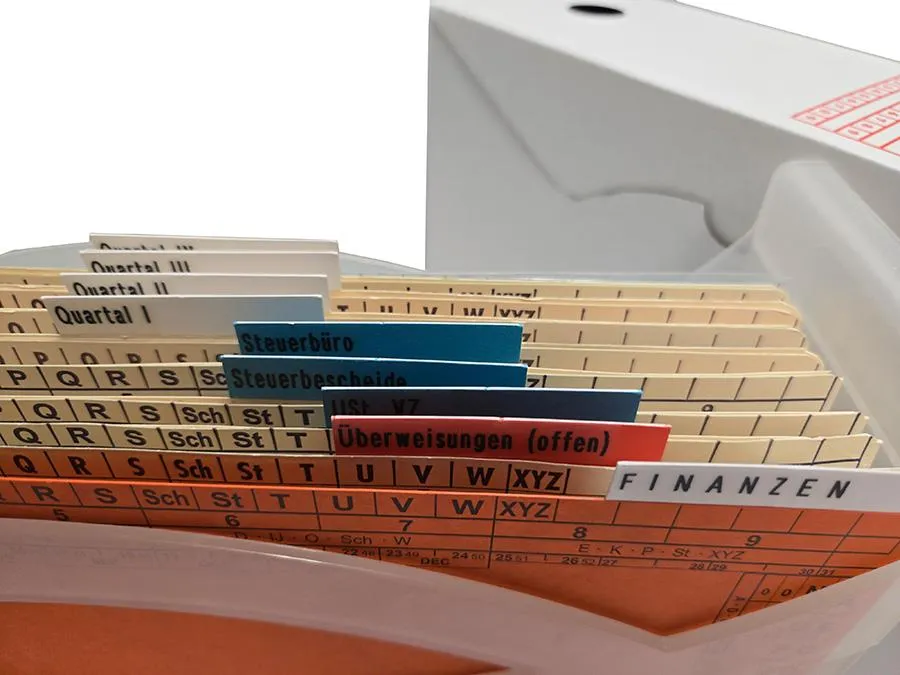 Nahaufnahme einer Mappei Ordnungsbox, vorne in der Box befindet sich eine Leitkarte, dahinter mehrere Ordnungsmappen mit angeklebten Taben. Hinter der Ordnungsbox befindet sich eine Archivschachtel